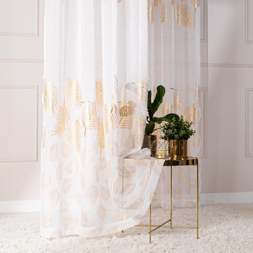 Biała, transparentna firana ze złotym nadrukiem w kształcie liści, opadająca do podłogi. Firana zawieszona jest na karniszu i delikatnie pofałdowana. Obok firany stoi mały, złoty stolik z doniczkami z zielonymi roślinami. W tle widoczna jest biała ściana 
