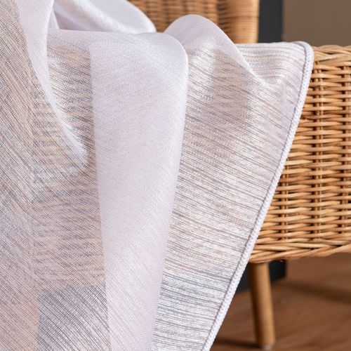 Zbliżenie na strukturę białej firany Sawina z pionowymi, błyszczącymi pasami, udrapowanej na wiklinowym oparciu.